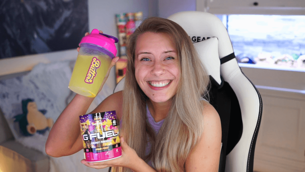 NoisyButters is holding her G FUEL Star Fruit shaker cup and tub while smiling and sitting in a gaming chair.